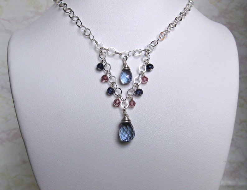 May include: A silver chain necklace with a teardrop-shaped blue gemstone pendant. The pendant is surrounded by smaller blue and pink gemstones.