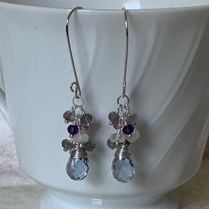 May include: Silver dangle earrings with a teardrop shaped blue gemstone and smaller stones in shades of gray, pink, and purple.