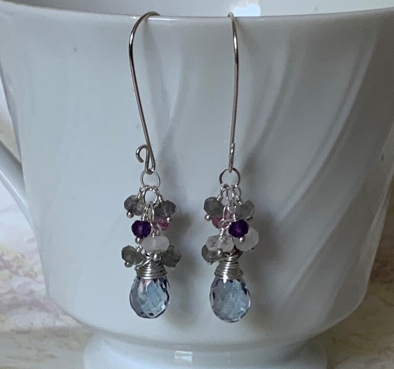 May include: Silver dangle earrings with a teardrop shaped blue gemstone and smaller stones in shades of gray, pink, and purple.