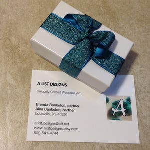 May include: A white gift box with a teal blue ribbon tied in a bow. The box is sitting on a business card with the text "A List Designs" and contact information for Brenda and Alea Bankston.