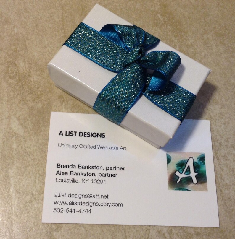 May include: A white gift box with a teal blue ribbon tied in a bow. The box is sitting on a business card with the text "A List Designs" and contact information for Brenda and Alea Bankston.