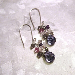 May include: A pair of silver dangle earrings with a cluster of purple, pink, and white gemstones. The earrings feature a teardrop-shaped purple gemstone at the bottom.