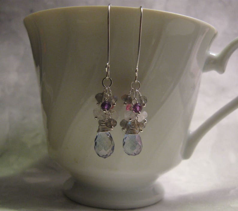 May include: A pair of silver earrings with a cluster of light blue, purple, and pink gemstones. The earrings are hanging from a white teacup.