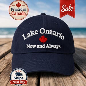 May include: A navy blue baseball cap featuring white embroidered text that reads Lake Ontario Now and Always with a red maple leaf icon centred between the lines of text. The cap has a structured crown and a curved brim.