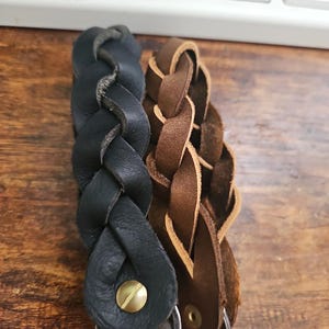 Op de afbeelding: Twee gevlochten leren sleutelhangers, een zwarte en een bruine, met metalen sleutelringen.