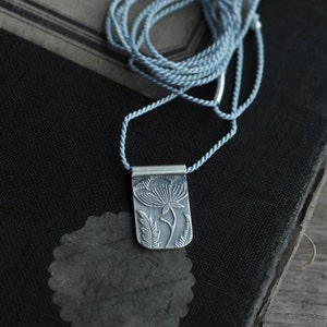 May include: A silver necklace with a delicate floral pendant. The pendant is a rectangular shape with a detailed floral design. The necklace is on a blue cord.
