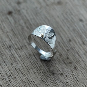 May include: A silver ring with a detailed design of leaves and a small gold dot in the center.