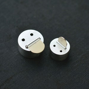 May include: Two silver metal round objects with a small flap on top. The larger object has a gold colored flap.