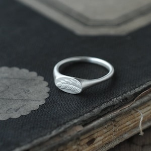 May include: A silver signet ring with a leaf design on a black book.