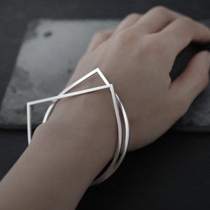 May include: Two silver geometric bracelets stacked on a wrist. The bracelets are made of thin metal and have a simple, modern design.