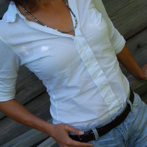 May include: A white button-down shirt with a single pocket, paired with light blue jeans and a dark brown belt. A beaded necklace adds a touch of color. The model is leaning against a wooden structure.