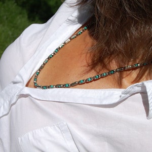 May include: A silver necklace with turquoise-colored stones. The necklace is composed of small, rectangular silver links alternating with the turquoise stones. The necklace is worn over a white collared shirt.