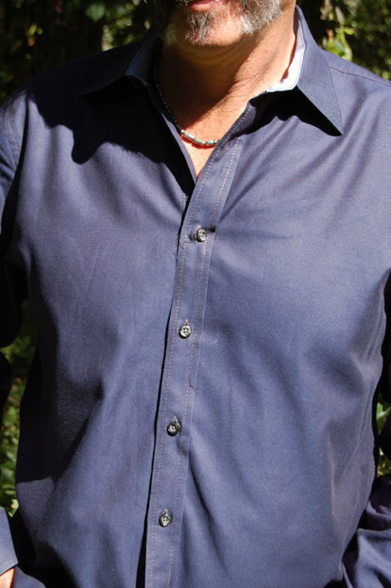 May include: A navy blue button-down shirt with a white collar and light brown stitching. The shirt has dark buttons and is partially unbuttoned, revealing a silver necklace. The fabric appears to be a smooth, woven material.