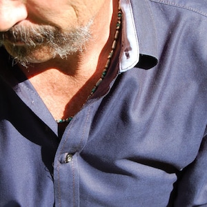 May include: A close-up of a navy blue button-down shirt with a white and light blue collar. A beaded necklace with turquoise and tan beads is visible. The shirt is buttoned at the top.