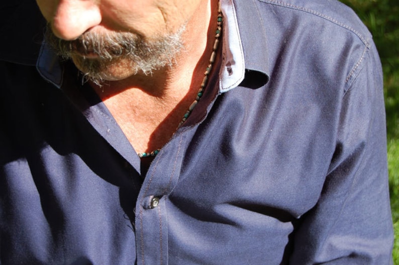May include: A close-up of a navy blue button-down shirt with a white and light blue collar. A beaded necklace with turquoise and tan beads is visible. The shirt is buttoned at the top.