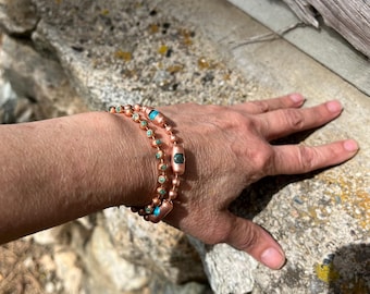 May include: A double-strand bracelet featuring copper-toned metal beads and small, round, turquoise-colored stone accents. The design incorporates cylindrical copper beads interspersed with smaller spherical beads, creating a layered appearance.