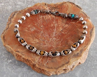 May include: A silver bracelet with brown tiger's eye beads and turquoise accents. The bracelet is on a brown wooden surface.