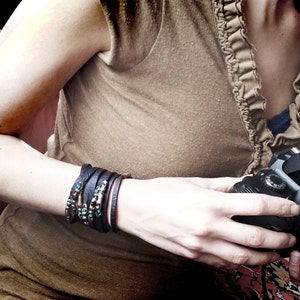 以下が含まれることがあります： 茶色のフリル付きトップスを着て、手首に複数の革のブレスレットをしている女性が、フラッシュが取り付けられた黒いカメラを持っている。