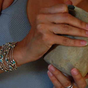 May include: A woman's hand holds a brown clay pot with a rough texture. The woman is wearing a gray tank top and a bracelet with turquoise and gold beads.