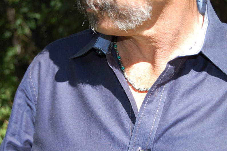 May include: A close-up of a navy blue button-down shirt with a contrasting light blue collar and cuffs. The shirt has white stitching and is paired with a silver and turquoise beaded necklace. The image is taken outdoors.