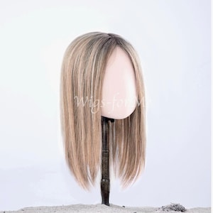 Peut inclure: Une perruque mi-longue aux cheveux blonds raides et nuancés, avec des racines plus foncées et des mèches plus claires. La perruque est présentée sur un support sombre sur fond clair, avec le texte Wigs-for-Me visible sur la chevelure.