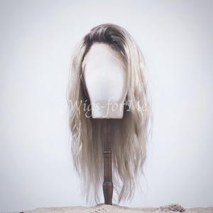 Puede incluir: Una peluca larga y ondulada, de color rubio claro, con una raya natural. La peluca está en una cabeza de maniquí y se muestra sobre un fondo blanco.