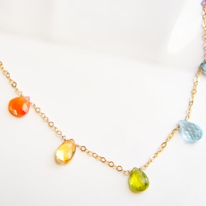 Könnte beinhalten: Eine zarte goldene Halskette mit tropfenförmigen Edelsteinen in Regenbogenfarben. Die Edelsteine umfassen Rot, Orange, Gelb, Grün, Blau und Lila. Die Halskette wird auf einer weißen Oberfläche präsentiert.