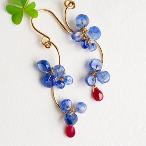 May include: A pair of gold-toned wire earrings featuring clusters of blue gemstones and a single red gemstone drop. The earrings have a curved design and are displayed on a white background. A green shamrock is in the upper left corner.
