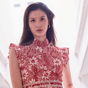 Half body view of young Eurasian  model, with gold earrings and red white batik dress. Dress has a stand up mandarin collar, and pleated trimming which looked like sleeves. There is also a waistband with lace trimming.