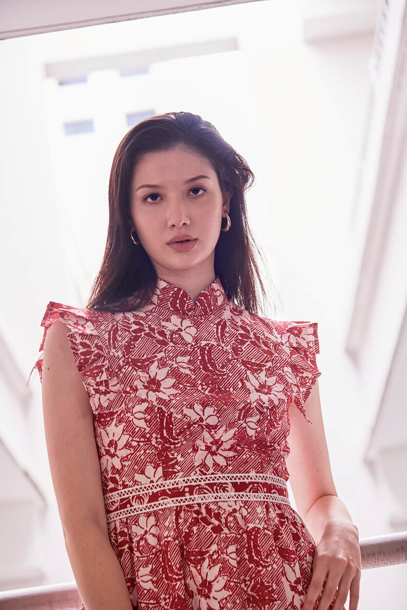 Half body view of young Eurasian  model, with gold earrings and red white batik dress. Dress has a stand up mandarin collar, and pleated trimming which looked like sleeves. There is also a waistband with lace trimming.