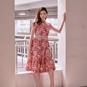 Young Eurasian lady in red and white batik cheongsam dress, standing with her arm on the wall. Dress is mid length and has a lace trimmed waist band.