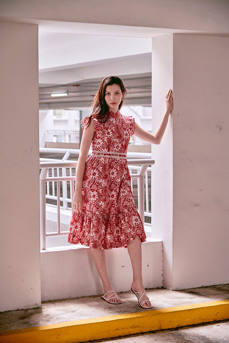 Young Eurasian lady in red and white batik cheongsam dress, standing with her arm on the wall. Dress is mid length and has a lace trimmed waist band.