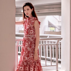 Side profile of Eurasian model, she is modelling a Dayglow red and white batik qipao dress. The dress is over her knee and has a frilly hem and laced trimmed waist band.