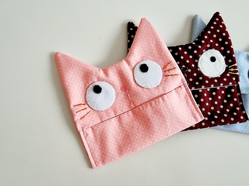 May include: Three pink, brown, and blue fabric cat-shaped pouches with white polka dots. Each pouch has embroidered whiskers and white felt eyes with black pupils.