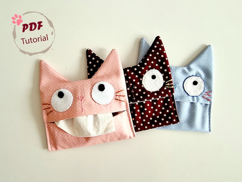 May include: Three cat-shaped tissue box covers. The covers are made of fabric with polka dots. The pink cover has a white tissue inside. The brown cover has white polka dots on a dark brown background. The blue cover has white polka dots on a light blue background.