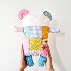 May include: A colourful patchwork bear cushion. The bear has a white head, pink and blue ears, and a body made of various fabric squares in pastel shades and floral designs. The bear's arms and feet are also made of patterned fabric.