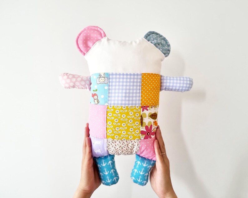 May include: A colourful patchwork bear cushion. The bear has a white head, pink and blue ears, and a body made of various fabric squares in pastel shades and floral designs. The bear's arms and feet are also made of patterned fabric.