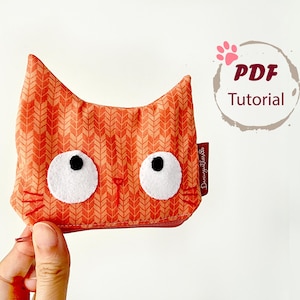 May include: Orange cat-shaped zippered pouch with white felt eyes and whiskers. The pouch has a patterned design and a small red tag. The image includes the text "PDF Tutorial" in a circular design.