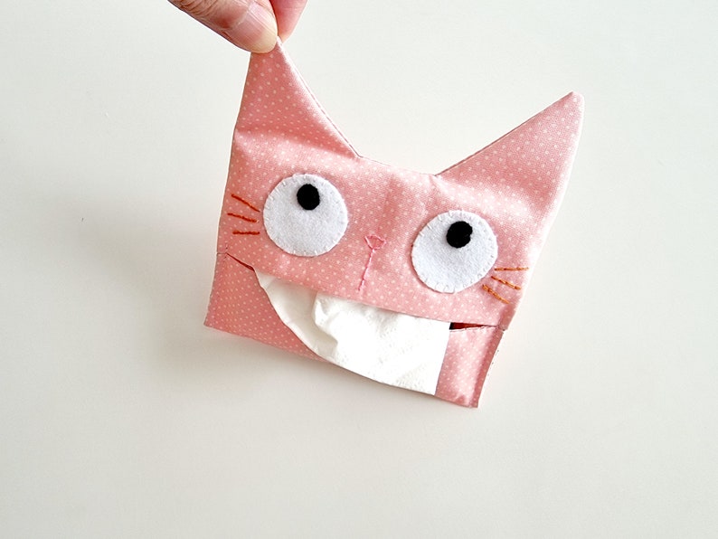 May include: Pink fabric tissue box cover shaped like a cat with white eyes, black pupils, and orange whiskers.