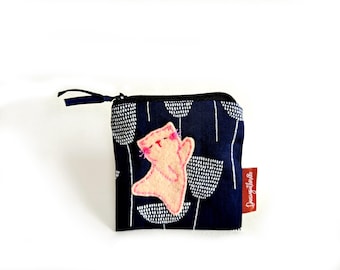 May include: A dark blue zippered pouch featuring a white abstract floral pattern and a light pink felt cat appliqué. The cat has embroidered pink facial features and whiskers. A small red fabric tag with white text is attached to the side.