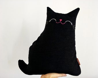 May include: A black cat-shaped pillow featuring embroidered white eyes and whiskers with a pink stitched nose. The fabric has a subtle textured appearance, and a small brown rectangular tag is visible on the lower right side of the cushion.