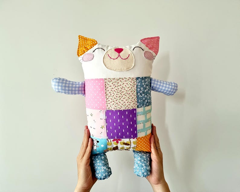May include: A handmade patchwork cat plush toy. It has a white face, pink and yellow ears, and a smiling face. The body is made of colourful fabric squares in various patterns and colours, with blue and pink gingham arms and blue patterned legs.