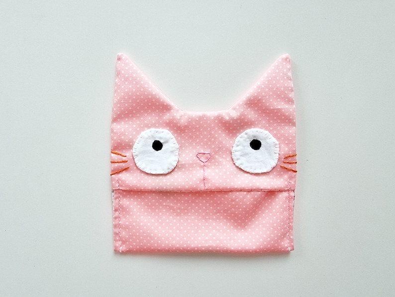 May include: A pink fabric cat-shaped pouch with white polka dots and embroidered white eyes with black pupils. The pouch has a pink nose and orange whiskers.