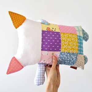 May include: A handmade patchwork goose-shaped pillow. The pillow has a white body, orange and pink accents, and a colourful patchwork design with various patterns and colours, including purple, yellow, and blue. The pillow is held by a hand.