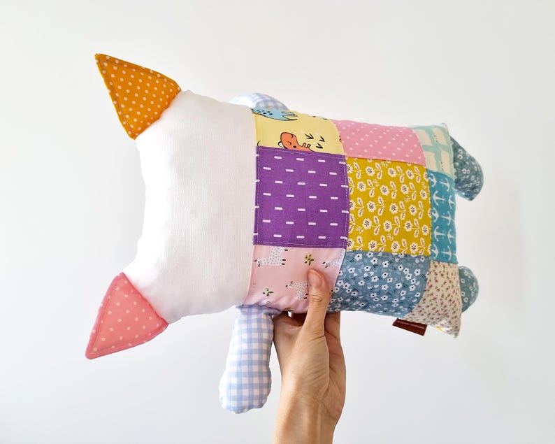 May include: A handmade patchwork goose-shaped pillow. The pillow has a white body, orange and pink accents, and a colourful patchwork design with various patterns and colours, including purple, yellow, and blue. The pillow is held by a hand.