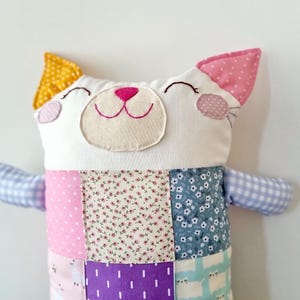 May include: A handmade cat-shaped soft toy with a patchwork design. The cat has a white face with embroidered features, a pink nose, and smiling eyes. The ears are yellow and pink. The body is made of various patterned fabrics in pink, purple, and blue.