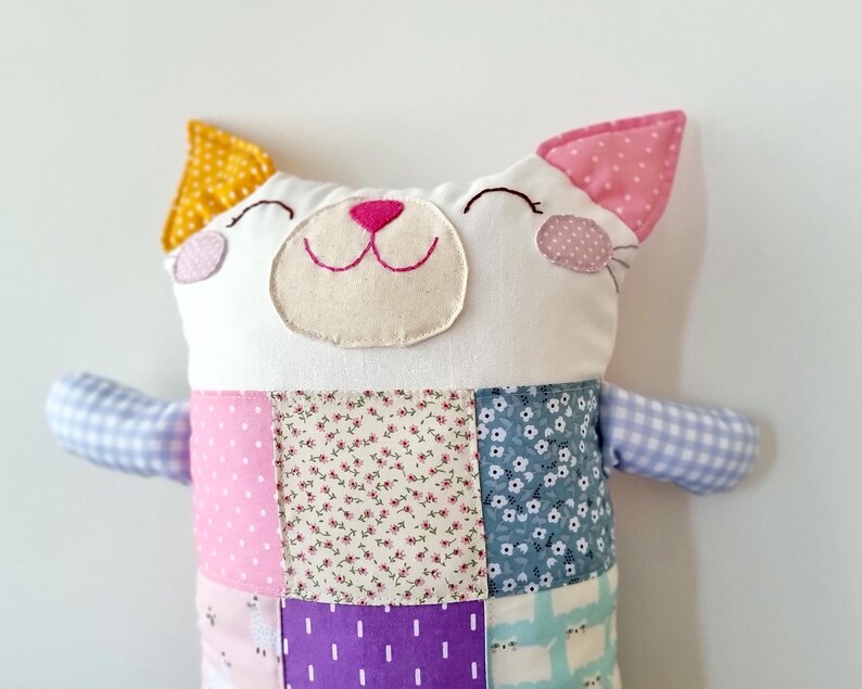 May include: A handmade cat-shaped soft toy with a patchwork design. The cat has a white face with embroidered features, a pink nose, and smiling eyes. The ears are yellow and pink. The body is made of various patterned fabrics in pink, purple, and blue.