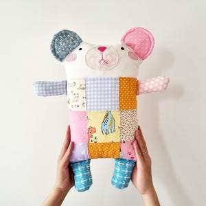 May include: A handmade patchwork bear plush toy with a white face, pink ears, and a patchwork body. The body features squares of various fabrics in colors like blue, orange, pink, and yellow. The bear's arms and legs are also made of patterned fabric.