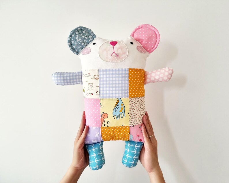 May include: A handmade patchwork bear plush toy with a white face, pink ears, and a patchwork body. The body features squares of various fabrics in colours like blue, orange, pink, and yellow. The bear's arms and legs are also made of patterned fabric.