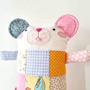 May include: A handmade stuffed bear with a patchwork body. The bear has a white face with pink accents, a pink nose, and two ears, one blue with white polka dots and the other pink with white polka dots. The bear's body is made of various patterned fabrics.